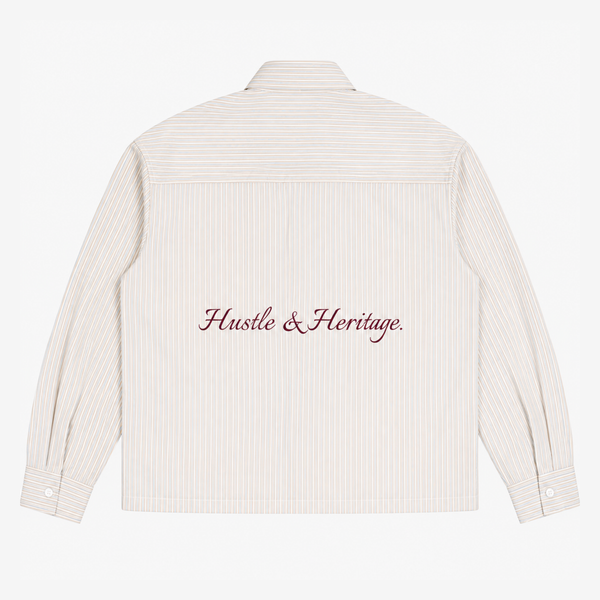 The back of a cream-colored long-sleeve shirt with thin vertical pinstripes and 'Hustle & Heritage.' embroidered in dark red script across the center.
